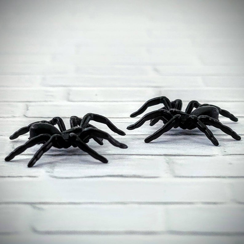 May include: Two black plastic spiders on a white brick background. The spiders have eight legs and are facing different directions.