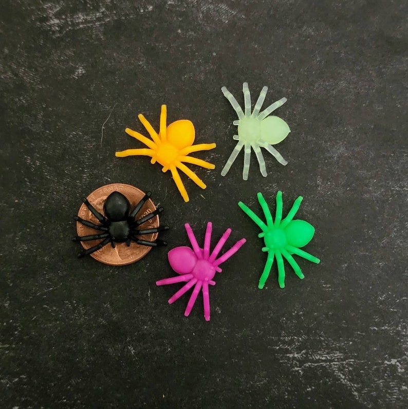 May include: Five plastic spiders in different colors: black, yellow, green, pink, and light green. The spiders are arranged in a circle on a dark gray background.