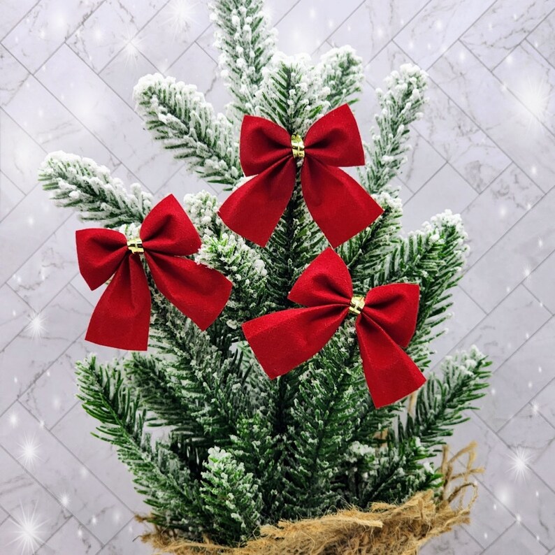 May include: Three red velvet bows with gold centres are attached to a small, snow-frosted artificial Christmas tree.