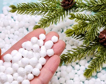 May include: A hand holding white, round decorative balls, with more scattered in the background. Green pine branches and brown pine cones are in the upper right corner, suggesting a holiday theme.