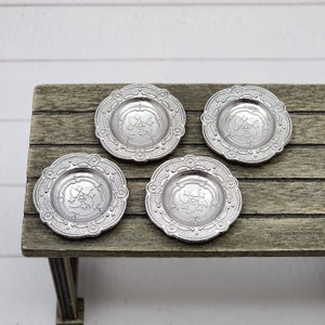 May include: Four silver decorative plates with an ornate design on a wooden table. The plates have a raised floral pattern in the center.