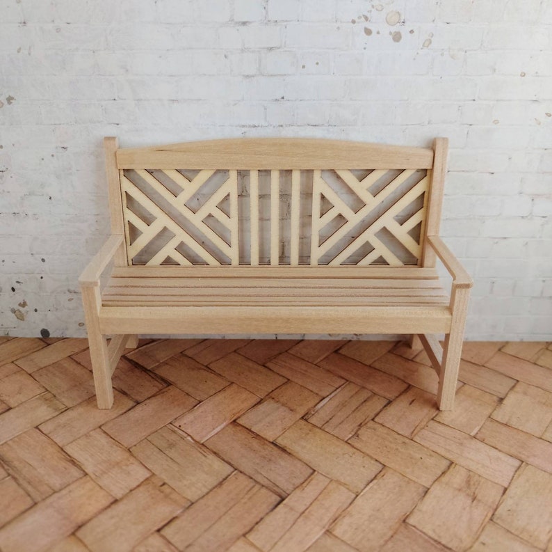 Op de afbeelding: Een houten bank met een geometrisch patroon op de rugleuning en een zitting met latten. De bank is lichtbruin en staat op een houten vloer.