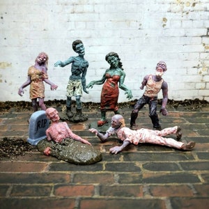 May include: A set of six zombie figurines, painted in shades of blue, green, and red. The figurines are posed in various positions, including standing, walking, and crawling. One zombie is lying on its back, partially buried in the ground, with a tombstone that reads "RIP" next to it.