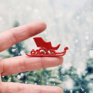 May include: A small, bright red miniature sleigh with curved runners and a seat. The sleigh is a festive decoration, perfect for holiday crafts or seasonal displays. The sleigh is approximately 1.5 inches long.