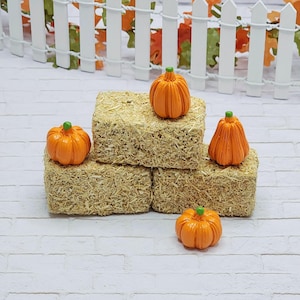 May include: Miniature orange pumpkins with green stems sit atop small hay bales. The pumpkins are arranged in a decorative display, perfect for fall or Halloween-themed crafts and decor. The background includes a white picket fence and autumn leaves.