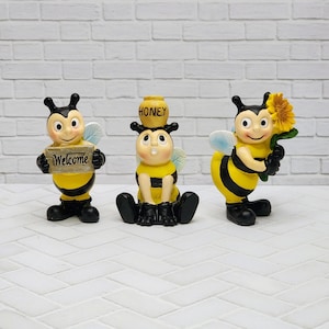 May include: Three whimsical bee figurines. One holds a "Welcome" sign, another balances a honey pot, and the third carries a sunflower. Each bee is yellow and black with black shoes and wings.