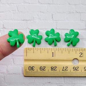 May include: Four green shamrock-shaped buttons, each measuring approximately 1 inch in diameter, are arranged in a row on a wooden ruler. The ruler is marked with inches and centimeters.