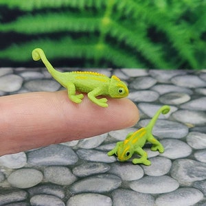 May include: Two small green plastic toy chameleons with yellow stripes. The chameleons are posed on a finger and a stone surface.