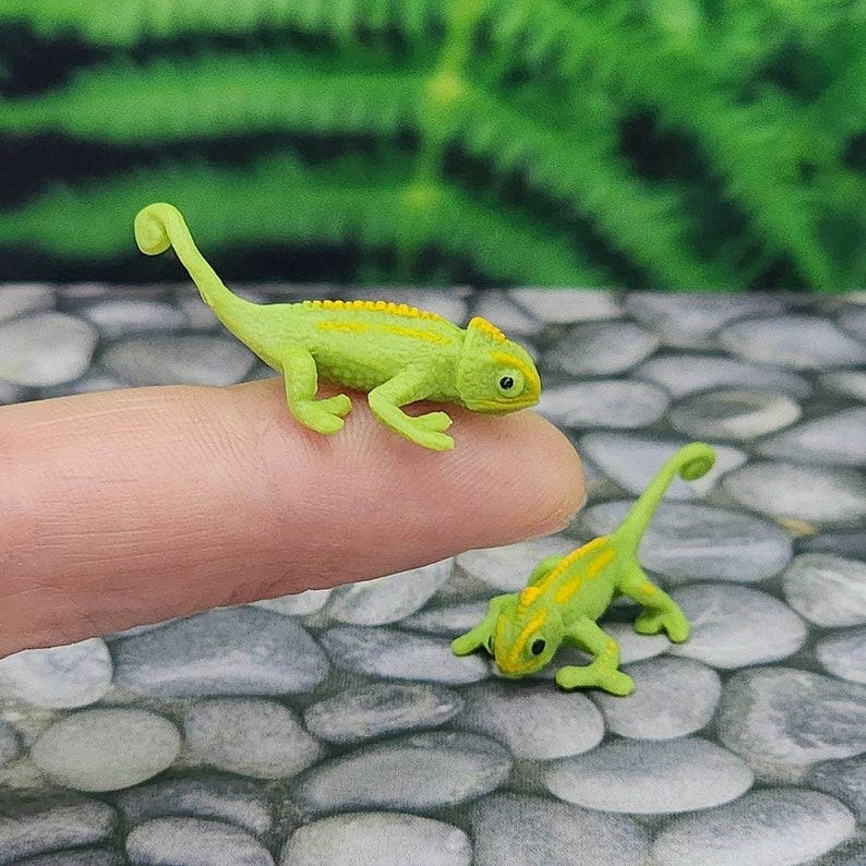 May include: Two small green plastic toy chameleons with yellow stripes. The chameleons are posed on a finger and a stone surface.
