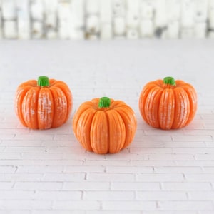 May include: Three miniature orange pumpkins with green stems. The pumpkins are arranged in a row on a white brick surface.