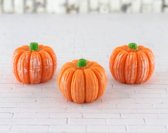 Puede incluir: Tres calabazas naranja en miniatura con tallos verdes. Las calabazas están dispuestas en fila sobre una superficie de ladrillo blanco.
