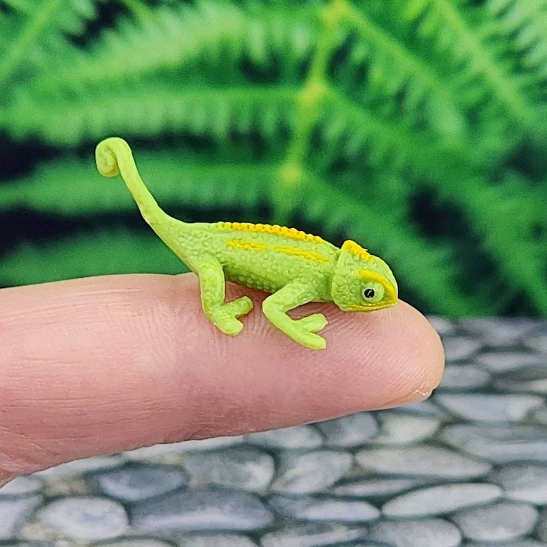 May include: A small green chameleon figurine with yellow stripes, posed on a finger.