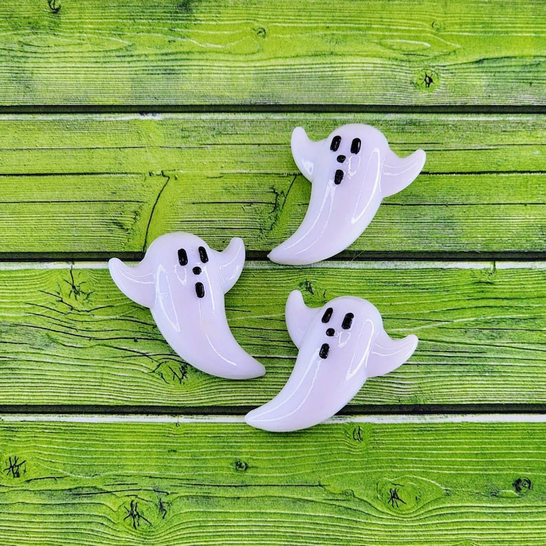 May include: Three white ghost-shaped buttons with black eyes and mouths. The buttons are on a green wooden surface.