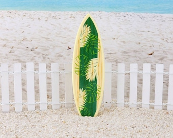 May include: A wooden surfboard with a green and white tropical leaf design. The surfboard is standing upright on a white picket fence in front of a sandy beach.