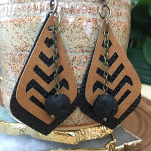 Puede incluir: Un par de pendientes de cuero marrón con recortes geométricos negros y cuentas de lava negra. Los pendientes tienen una cadena de latón que cuelga de la parte superior.