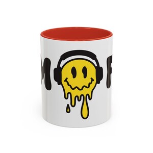 May include: White ceramic mug with a red rim. The mug features a black and yellow smiley face with headphones, dripping yellow paint. The text "Mood" is printed on either side of the smiley face.