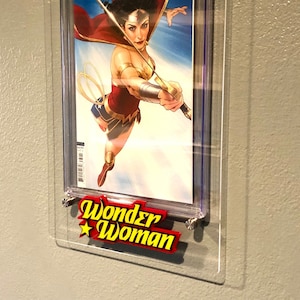 May include: A graded comic book featuring Wonder Woman in a red and gold costume, holding a sword and shield. The comic book is encased in a clear plastic case with a 9.8 rating label. The case is mounted on a wall with a Wonder Woman logo.