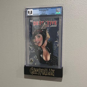 May include: A graded comic book featuring Catwoman from Batman Detective Comics #1000, encased in a clear protective holder. The holder is mounted on a black display stand with "Catwoman" in gold lettering.