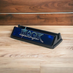 May include: A black rectangular card holder with the word "MAGIK" in white and blue lettering. The lettering is on a blue background. The holder is made of a black material and sits on a wooden surface.