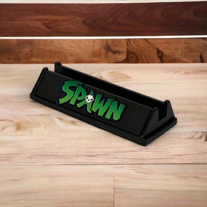 May include: Black desk organizer with the word "SPAWN" in green and blue lettering, featuring a skull graphic. The organizer is made of a glossy material and has a rectangular shape with angled sides.