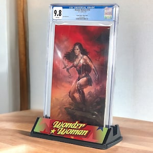 Puede incluir: Un cómic Wonder Woman #750 calificado y encapsulado, exhibido en un soporte con el logotipo de Wonder Woman. El cómic muestra a Wonder Woman con una espada sobre un fondo rojo. El soporte es negro.