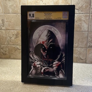 Puede incluir: Una portada de cómic en blanco y negro con un personaje con un disfraz con capucha y la cara ensangrentada. La portada está calificada con un 9,8 y está en una placa negra CGC Signature Series. El cómic se titula "Moon Knight #1".