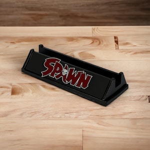May include: A black desk organizer with the word "Spawn" in red and white lettering. The organizer has a rectangular shape with a slot for holding items. The design includes a skull graphic.