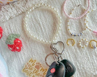 May include: A white pearl keychain with a heart charm and a key fob with a "Mom to the best pup" sticker. The keychain is on a white fluffy surface with other beaded jewellery.