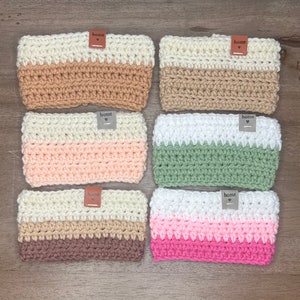 May include: Six crocheted cup cozies in various colour combinations. Each cozy has a white top band, a second band in a solid colour, and a third band in a different solid colour. Each cozy has a brown leather tag with the word "home" on it.