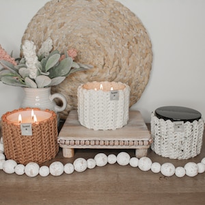 May include: Three crocheted candle jars with the word "home" on each. The candles are lit and the jars are on a wooden stand with white beads in front of them.