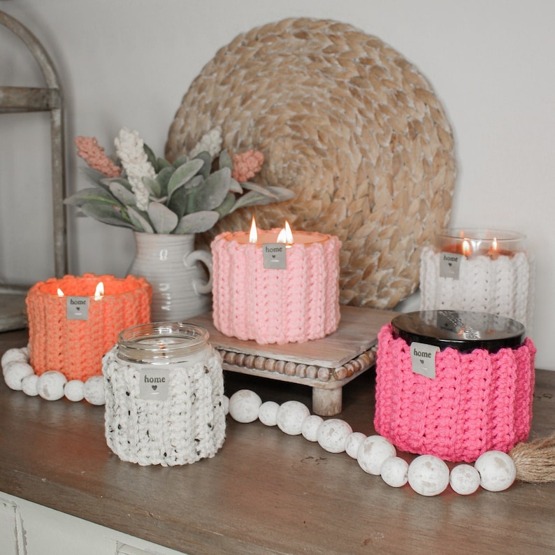 May include: Four candles in glass jars with crocheted covers in pink, white, and orange. The candles are lit and the covers have the word "home" on them.