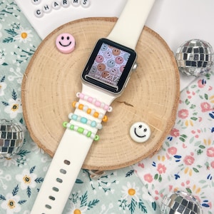 May include: White Apple Watch with a white band and a colorful beaded bracelet. The watch face displays a pattern of smiling faces. The watch is on a wooden surface with a pink and white smiley face charm and a silver disco ball.