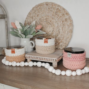May include: Three crocheted candle jars with lids. The jars are in shades of white, beige, and pink. The jars are sitting on a wooden tray with a woven mat underneath. The jars have the word "home" on them.