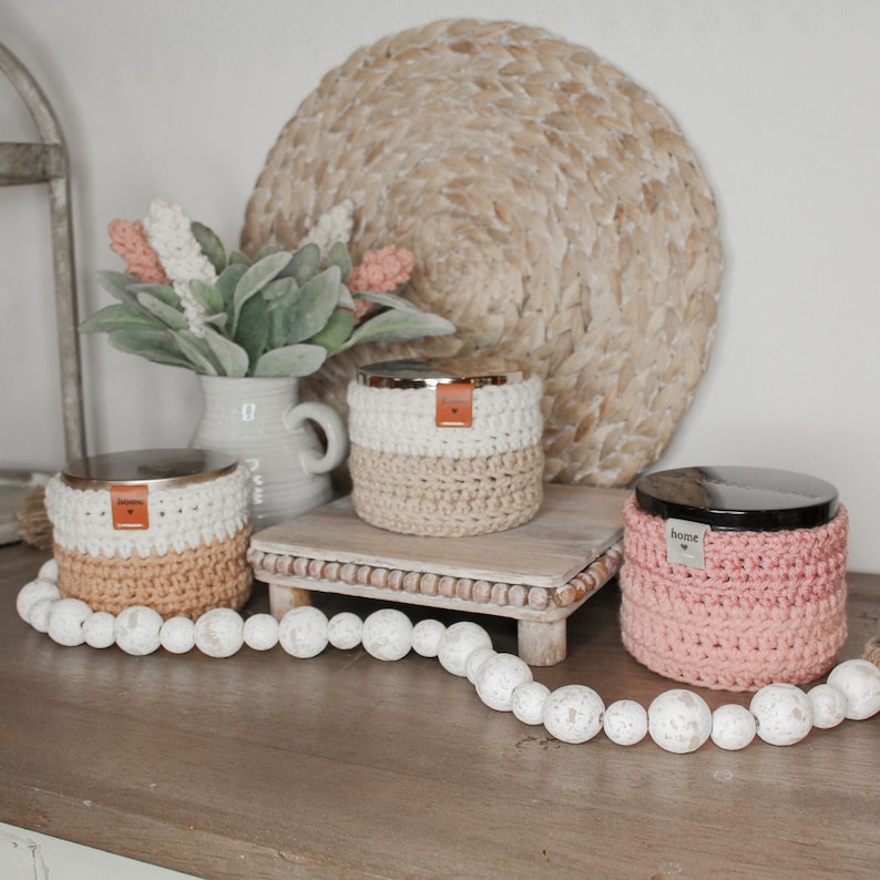 May include: Three crocheted candle jars with lids. The jars are in shades of white, beige, and pink. The jars are sitting on a wooden tray with a woven mat underneath. The jars have the word "home" on them.