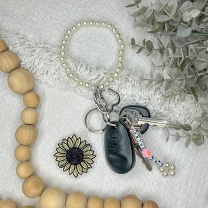 May include: A collection of accessories including a pearl bracelet, keys, and a sunflower keychain. The keys have a tag that says "DRIVE SAFE" with a pink flower. A wooden bead garland is in the foreground.