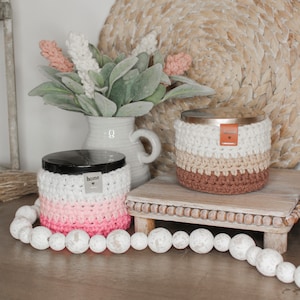May include: Two crocheted candle jars with lids. The jar on the left is pink and white and says "home" on the lid. The jar on the right is brown, beige, and white. Both jars are sitting on a wooden tray.