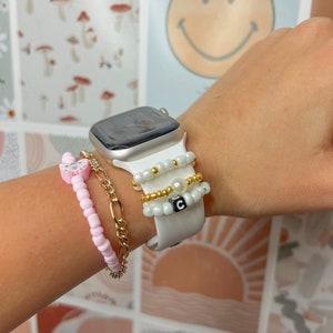 May include: A white Apple Watch with a white band and three beaded bracelets. The bracelets are white, gold, and pink with a small butterfly charm. The watch face is black and shows the time.