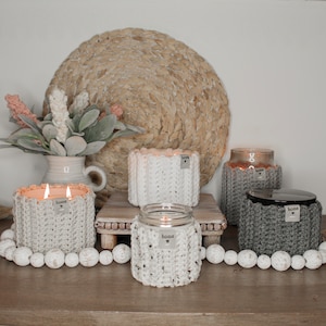 May include: Four candles in crocheted jars with the word "home" on each jar. The candles are lit and sitting on a wooden table with a woven mat and white beads.