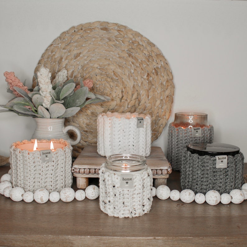 May include: Four candles in crocheted jars with the word "home" on each jar. The candles are lit and sitting on a wooden table with a woven mat and white beads.