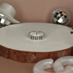 May include: A silver ring with the word "HARLOW" engraved on it. The ring is sitting on a wooden surface.