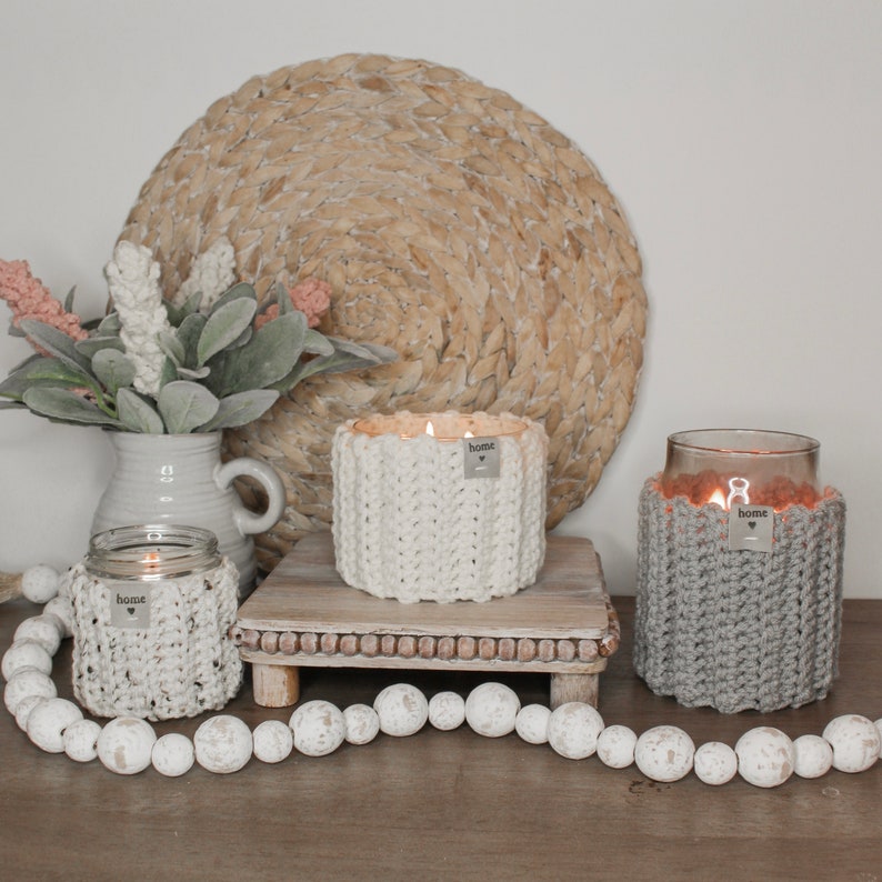 May include: Three crocheted candle jars with the word "home" on each. The jars are lit and sitting on a wooden tray with white beads around the base.