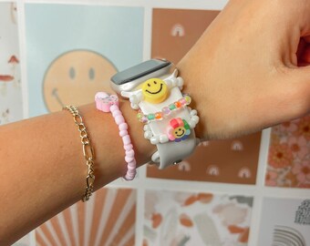 May include: A white Apple Watch with a white band and a smiley face charm. The watch is adorned with colourful beaded bracelets, including pink, yellow and blue beads.