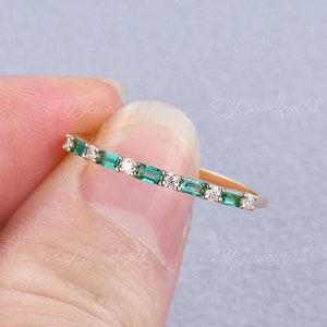 May include: A gold band ring featuring a row of emerald-cut green gemstones and small white gemstones. The ring is held by a person's hand against a light gray background. The text "TY JewelryUs" is visible in the background.