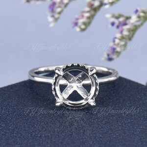 Puede incluir: Un engaste de anillo de plata con un centro redondo abierto para una piedra preciosa. El engaste tiene un diseño delicado e intrincado con pequeñas garras alrededor del borde.
