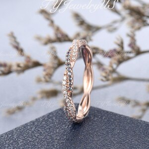 May include: Rose gold ring with a twisted design. The ring is encrusted with small, clear gemstones along the twisted band. The ring sits on a dark gray surface with a blurred floral background.
