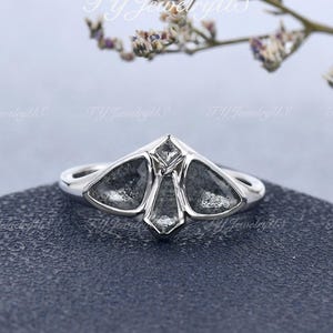 May include: A silver-toned ring featuring a moth-shaped centerpiece set with three triangular, speckled grey stones and one small, clear, diamond-shaped stone at the top. The band is smooth and polished, showcasing a symmetrical, winged design.