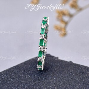 May include: A silver-toned eternity band featuring an alternating pattern of rectangular-cut green stones and round-cut clear stones. The ring is positioned vertically, displaying a continuous sequence of gemstones set in a metallic band.