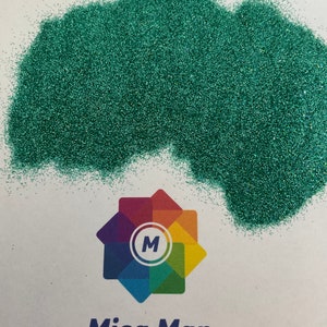 May include: A mound of shimmering teal glitter is displayed on a white background. The glitter is finely ground and sparkles. Beneath the glitter is a colourful logo with the text "Mica Man" in blue.