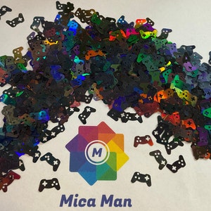 Puede incluir: Un montón grande de purpurina negra iridiscente y arcoíris con forma de mandos de videojuegos. La purpurina está esparcida sobre una superficie blanca con el texto "Mica Man" impreso debajo.