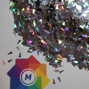 May include: A pile of iridescent silver glitter shaped like rockets. The glitter is scattered on a white surface with a rainbow star logo in the center.
